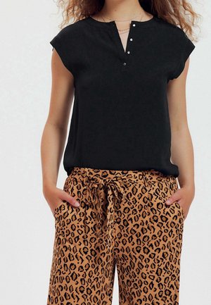 Femme portant un chemisier noir sans manches avec des boutons et un pantalon large imprimé léopard, les mains dans les poches.