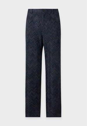 Junger Mann steht mit einer Hand in der Tasche, trägt ein leichtes kurzärmeliges Stricktop, dunkle gemusterte weite Hosen und schwarze Schuhe.