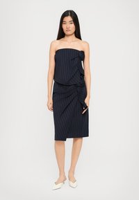 Vrouw in een strapless marineblauw gestreept jurkje met geraffineerde details en witte puntige hakken, staande tegen een effen achtergrond.