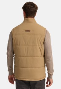 Tan puffer vest met een hoge kraag, voorzien van een horizontaal gestikt ontwerp en een leren accentlabel. Draagt over een grijze longsleeve shirt.