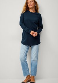 Marineblå langærmet top med rund halsudskæring, afslappet pasform og buet kant, som er sat sammen med lyseblå straight-leg jeans og sorte sandaler.