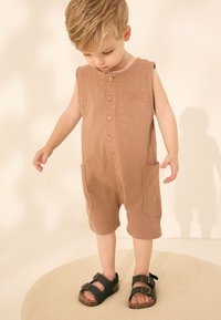 Brun, ærmeløs romper med knapper foran og sidelommer. Har et lille sol-logo. Kombineret med mørke sandaler med spænder.