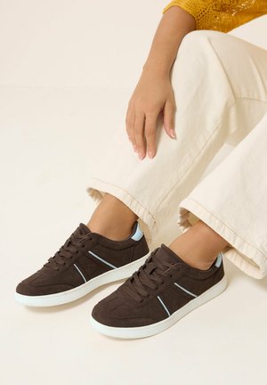 Personne portant des baskets brun foncé avec des semelles blanches et des accents bleu clair, assise jambes croisées, vêtue d'un pantalon crème et d'un haut jaune.