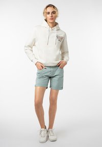 Crème-kleurige hoodie met een grafische print op de voorkant, lichtgroene shorts en witte sneakers. De capuchon heeft trekkoorden; de shorts hebben omgeslagen mouwen.