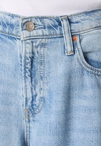 Jasnoniebieskie dżinsy z zapięciem na guzik, widocznymi szwami i kieszeniami z przodu. Tkanina wydaje się lekko wypłowiała i ma teksturowane wykończenie.