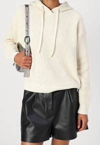 Felpa con cappuccio in maglia color crema con coulisse, abbinata a pantaloncini in pelle nera plissettati. L'outfit è completato da una borsa argento con dettaglio a tracolla.