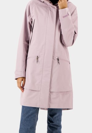 Kobieta ubrana w jasnoróżową, długą wodoodporną kurtkę z kieszeniami na suwak z przodu i niebieskie jeansy, trzymająca kołnierz kurtki jedną ręką.