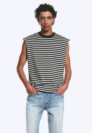 Homme aux cheveux bouclés portant un débardeur rayé noir et blanc et un jean bleu clair, les mains dans les poches, sur fond blanc.