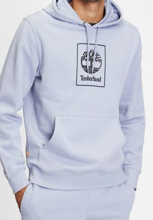 Hombre vistiendo una sudadera ligera de Timberland en color azul y pantalones a juego, con las manos en los bolsillos, logo de la sudadera visible y bolsillo frontal.