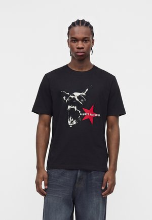 Mann mit geflochtenem Haar, der ein schwarzes T-Shirt mit einer Grafik eines knurrenden Hundes, einem roten Stern und dem Schriftzug „IMMER TREU“ trägt, kombiniert mit dunklen Jeans.