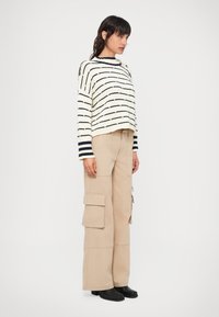 Meganinė juostuota megztinė smėlio spalvos su tamsiai mėlynomis linijomis, derinama su šviesiai smėlio spalvos kargo kelnėmis, turinčiomis kelis kišenių, ir juodais auliniais batais iki kulkšnies.