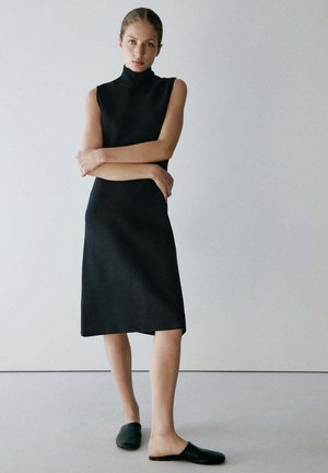 Femme debout, bras croisés, portant une robe midi noire sans manches et des chaussures noires sans lacets, sur un fond clair uni.