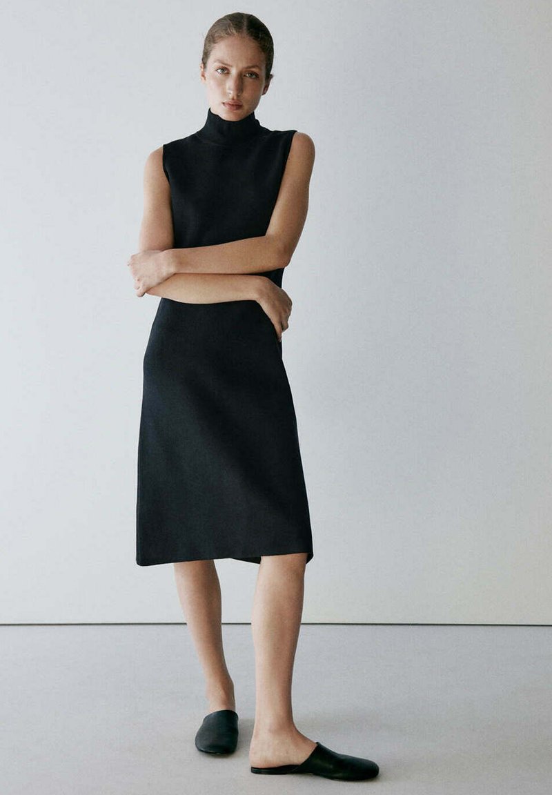 Femme debout, bras croisés, portant une robe midi noire sans manches et des chaussures noires sans lacets, sur un fond clair uni.