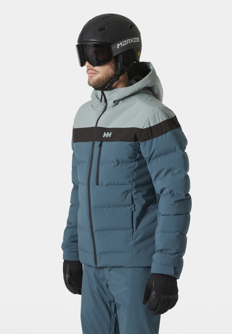 Veste de ski isolée bleue et grise avec une bande noire horizontale, capuche et poches zippées, portée avec des gants noirs et un casque.
