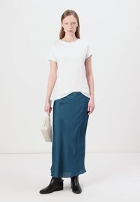 T-shirt bianco in cotone abbinato a una gonna lunga in satin color teal, con una vestibilità ampia e una leggera lucentezza. Stivali neri alla caviglia e una piccola borsa bianca.
