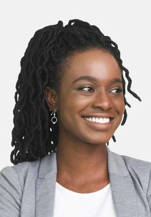 Una donna con capelli ricci scuri porta un blazer grigio chiaro sopra una maglietta bianca. Ha un sorriso discreto e orecchini a cerchio in argento.