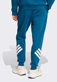 Turquoise joggers made of soft fabric, featuring white three-stripe accents on the legs. Ribbed cuffs and a comfortable fit at the waist.