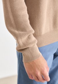 Beżowy sweter robiony na drutach z prążkowanymi mankietami i dołem, noszony z jasnoniebieskimi spodniami. Cechuje się gładką fakturą i dopasowanym krojem w okolicy nadgarstka.