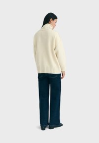 Elfenbeinfarbener, übergroßer Strickpullover mit hohem Kragen und gerippten Bündchen, kombiniert mit dunkelblauen weit geschnittenen Jeans. Weiche Textur und entspannte Passform.