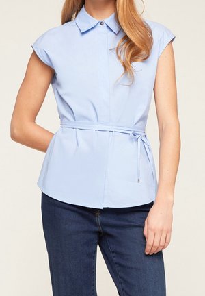 Femme portant un chemisier sans manches bleu clair à col avec ceinture à la taille et un jean bleu foncé, debout devant un fond uni.