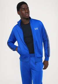 Man wearing a blue EA7 Emporio Armani tracksuit with black stripes on sleeves and a black shirt, looking to the side with hands in pockets.