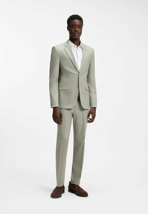 Egy férfi áll, világosszürke öltönyt, fehér inget és barna mokaszint viselve, egyszínű fehér háttér előtt.