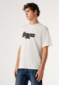 Biele bavlnené tričko s čiernym grafickým logom obsahujúcim nápis "73 PEPE JEANS LONDON." Model ho kombinuje s tmavomodrými denimovými džínsy.