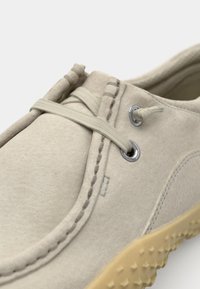 Beige semsket sko med rund tå, sømdetaljer, gummisåle og grå elastiske snørebånd festet til sølvfargede hemper.