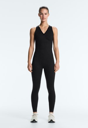 Donna in piedi di fronte, indossa una tuta nera aderente senza maniche e scarpe da ginnastica bianche su uno sfondo bianco.