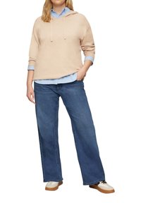 Helle beige Kapuzenpullover mit Kordelzug, getragen über einem hellblauen Button-up-Hemd, kombiniert mit weit geschnittenen blauen Jeans und hellbraunen Sneakern.