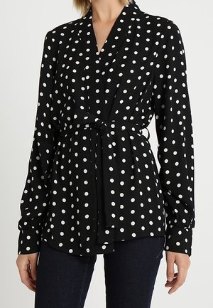 Zwarte blouse met lange mouwen en witte polka dots, met overslagstijl en een strik in de taille, gedragen met donkere broek.