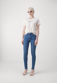 Striped t-skjorte i krem og lys gul med logo, kombinert med blå skinny jeans og klare hæler, båret av en modell med solbriller.