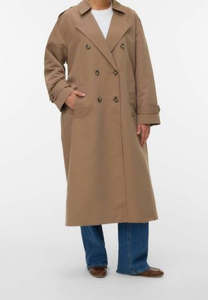 Trench-coat long beige à double boutonnage avec de larges revers porté sur une chemise blanche, un jean bleu et des chaussures marron, mains dans les poches.