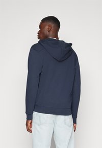 Marineblauwe hoodie met een gladde textuur, geribde manchetten en zoom. Heeft een kangoeroezak en stikselaccenten. Gemaakt met lichte spijkerbroeken.