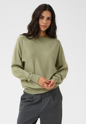 Femme aux longs cheveux foncés portant un pull vert clair à manches longues et un pantalon gris, debout, les mains jointes devant.