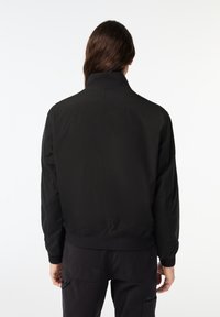 Homme aux longs cheveux bruns portant une veste noire à col montant et un pantalon noir, vu de dos sur un fond blanc.