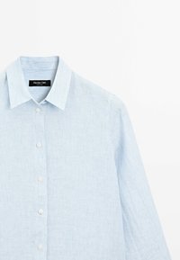 Chemise bleu clair à manches longues boutonnée avec des rayures verticales subtiles et un col pointu, présentée sur fond blanc.