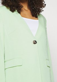 Jasnozielona marynarka z dekoltem w serek, teksturowanym materiałem oraz pojedynczym złotym zapięciem, posiadająca dwie przednie kieszenie.