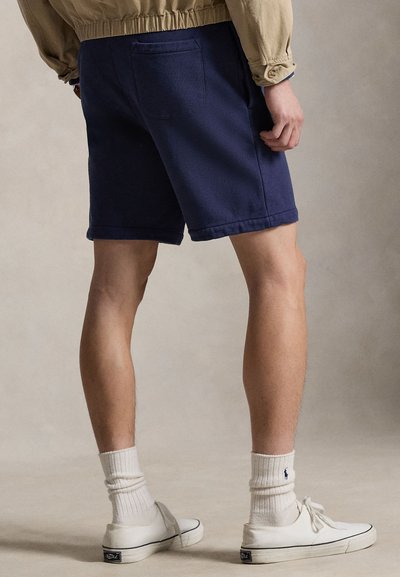Tamsiai mėlyni šortai iš medvilnės mišinio, su užpakaline kišene ir susuktomis apačiomis. Derinami su baltais sportbačiais ir raukšlėtomis kojinėmis.