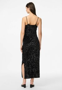 Crna maxi haljina s perlicama i tankim trakama, s bočnim prorezom i uskim siluetom, naglašava svoj sjajni izgled i dizajn.