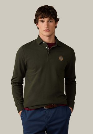 Jonge man draagt een donkergroen poloshirt met lange mouwen en een embleem, een blauwe broek en een bruine riem, staand met zijn handen in zijn zakken.