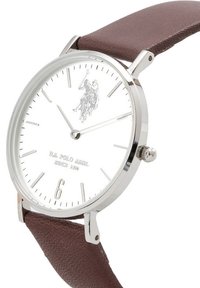 Orologio argento con quadrante bianco e cinturino in pelle marrone. Presenta marcatori delle ore minimalisti, un 6 in rilievo e un logo di un giocatore di polo.