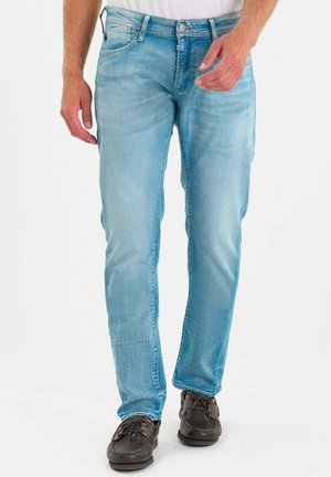 Man draagt lichtblauwe jeans en donkerbruine veterschoenen, staand tegen een witte achtergrond.