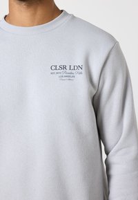 Hellgrauer Pullover mit geripptem Halsausschnitt, aus weicher Baumwolle. Verfügt über ein blaues Textlogo auf der linken Brustseite, mit Gründungsdatum und Standort.