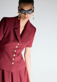 Donna che indossa un blazer su misura bordeaux con bottoni dorati e perle, una gonna abbinata, grandi orecchini dorati e con perle, e occhiali da sole a forma di occhio di gatto neri.