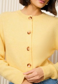 Gilet en tricot jaune avec un col rond, quatre grands boutons et des poignets côtelés. Texture douce et design ajusté.