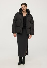 Giacca a vento nera con cappuccio, caratterizzata da un design trapuntato e due tasche frontali. Indossata sopra un top nero in maglia e una gonna nera elegante.