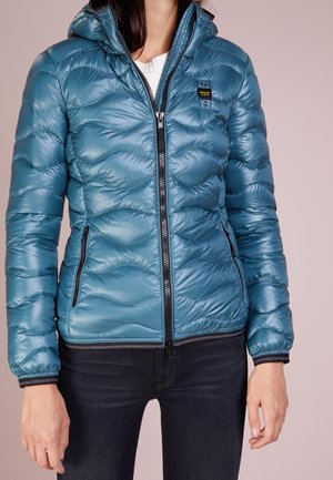 Femme portant une veste matelassée zippée brillante de couleur bleu sarcelle avec des détails de fermeture éclair noirs et un petit écusson logo sur la poitrine, associée à un jean foncé.