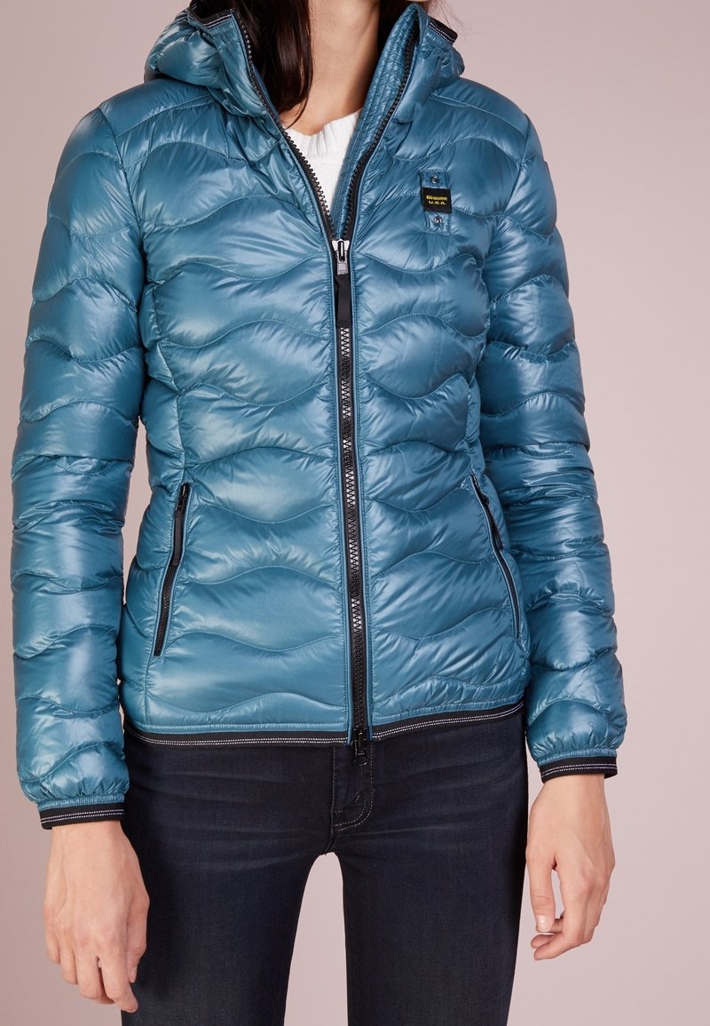 Femme portant une veste matelassée zippée brillante de couleur bleu sarcelle avec des détails de fermeture éclair noirs et un petit écusson logo sur la poitrine, associée à un jean foncé.