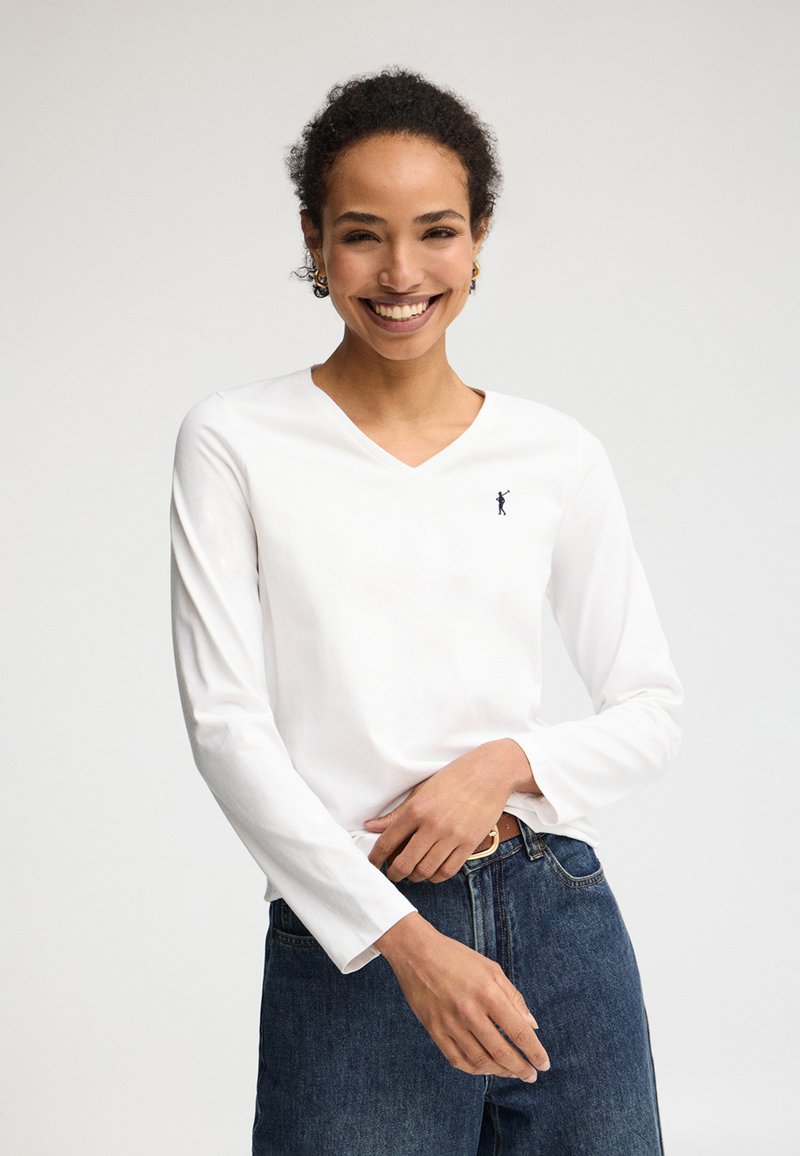 Glunderende vrouw met krullend haar, gekleed in een wit V-hals shirt met lange mouwen en een blauwe spijkerbroek, staand tegen een effen achtergrond.